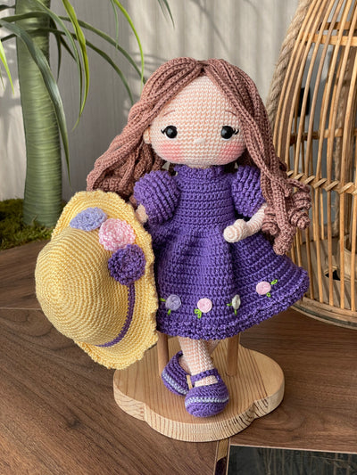 Handmade crocheted Purple Rose Girl Doll with long brown hair, wearing a purple dress with floral accents and a yellow hat. The doll is customizable with a name on a leather label, made using amigurumi techniques. Perfect gift for kids or room decor.