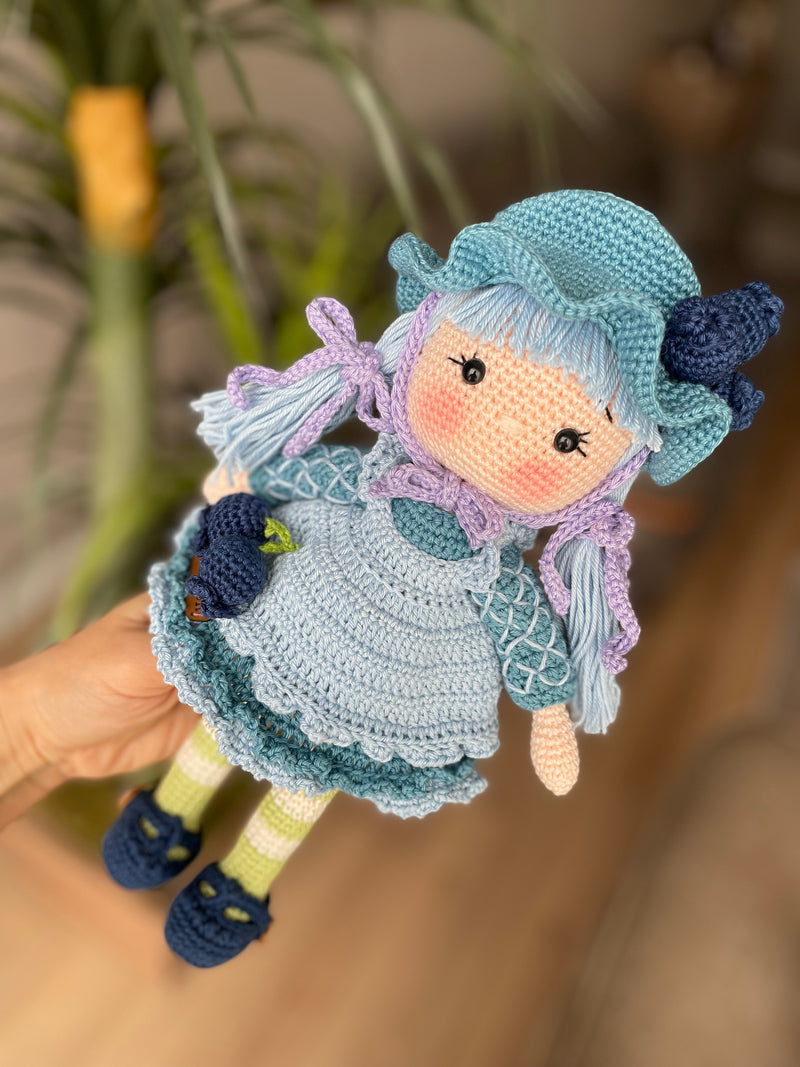 A handcrafted Blueberry Muffin Girl Doll featuring a detailed crochet blueberry accessory design, wearing a blue dress and hat, and personalized with a custom name on a leather label.