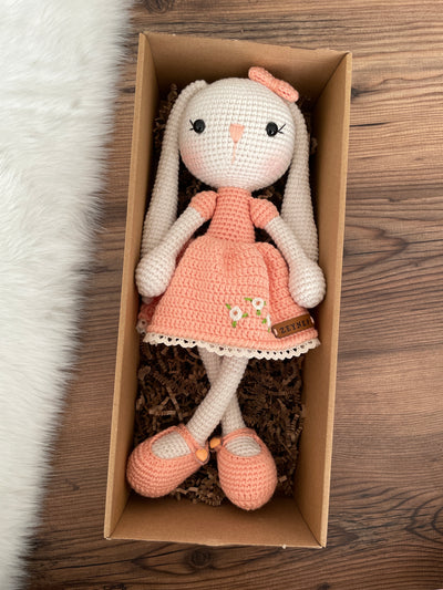 Handmade Salmon Dressed Bunny Rabbit Toy crocheted with amigurumi technique, featuring a cute salmon dress with embroidered flower details and customizable name on a leather label. Lying inside a gift box. Perfect for kids and baby gifts.