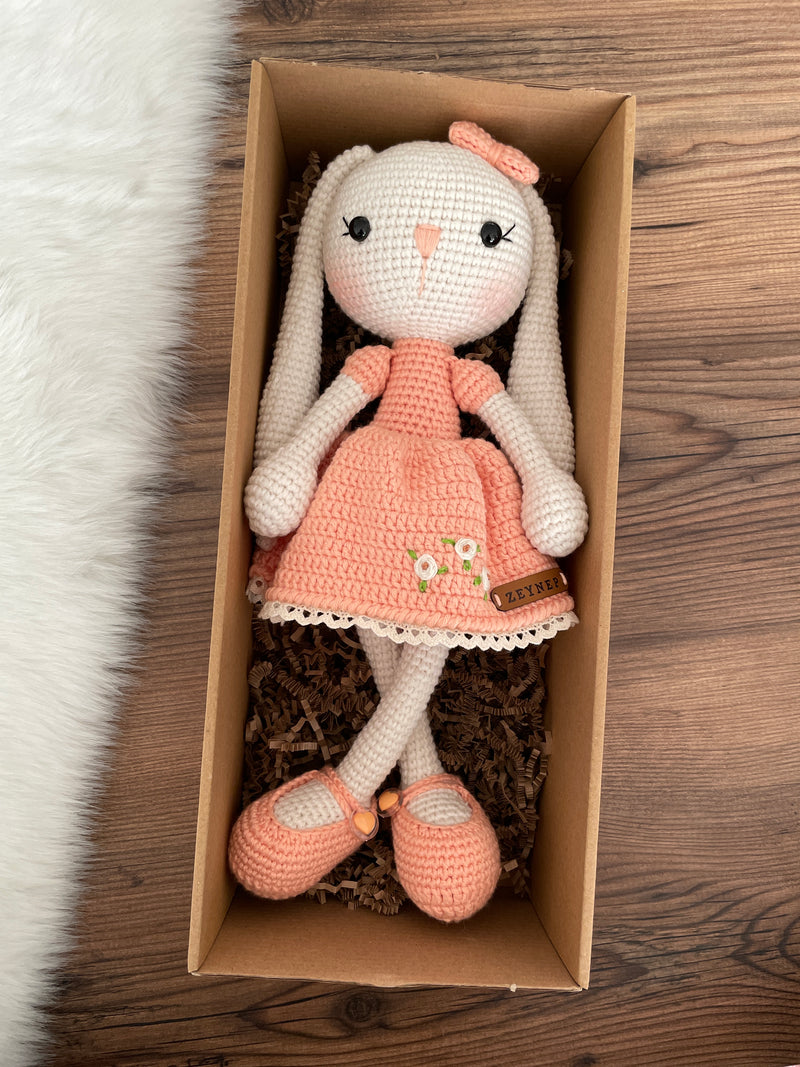 Handmade Salmon Dressed Bunny Rabbit Toy crocheted with amigurumi technique, featuring a cute salmon dress with embroidered flower details and customizable name on a leather label. Lying inside a gift box. Perfect for kids and baby gifts.