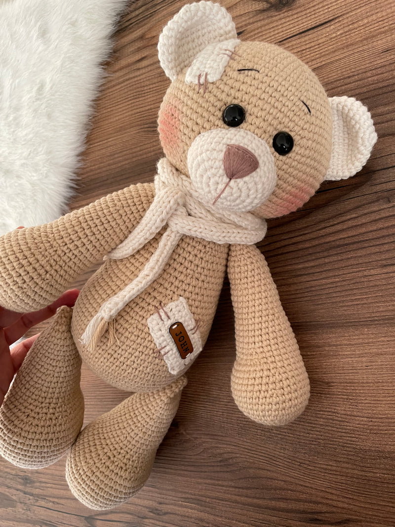 A handmade beige teddy bear toy crafted with the amigurumi technique, featuring a soft texture, a personalized leather name patch, and a cute, cuddly design.