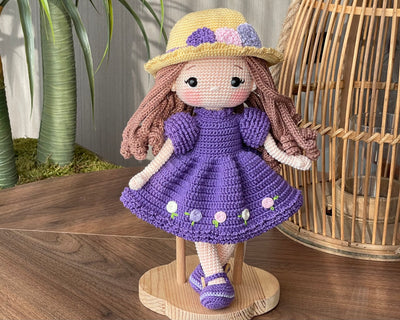 Handmade crocheted Purple Rose Girl Doll with long brown hair, wearing a purple dress with floral accents and a yellow hat. The doll is customizable with a name on a leather label, made using amigurumi techniques. Perfect gift for kids or room decor.