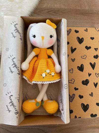 Handmade Yellow Dressed Bunny Rabbit Toy crocheted with amigurumi technique, featuring a cute yellow dress with embroidered flower details and customizable name on a leather label. Lying inside a gift box. Perfect for kids and baby gifts.