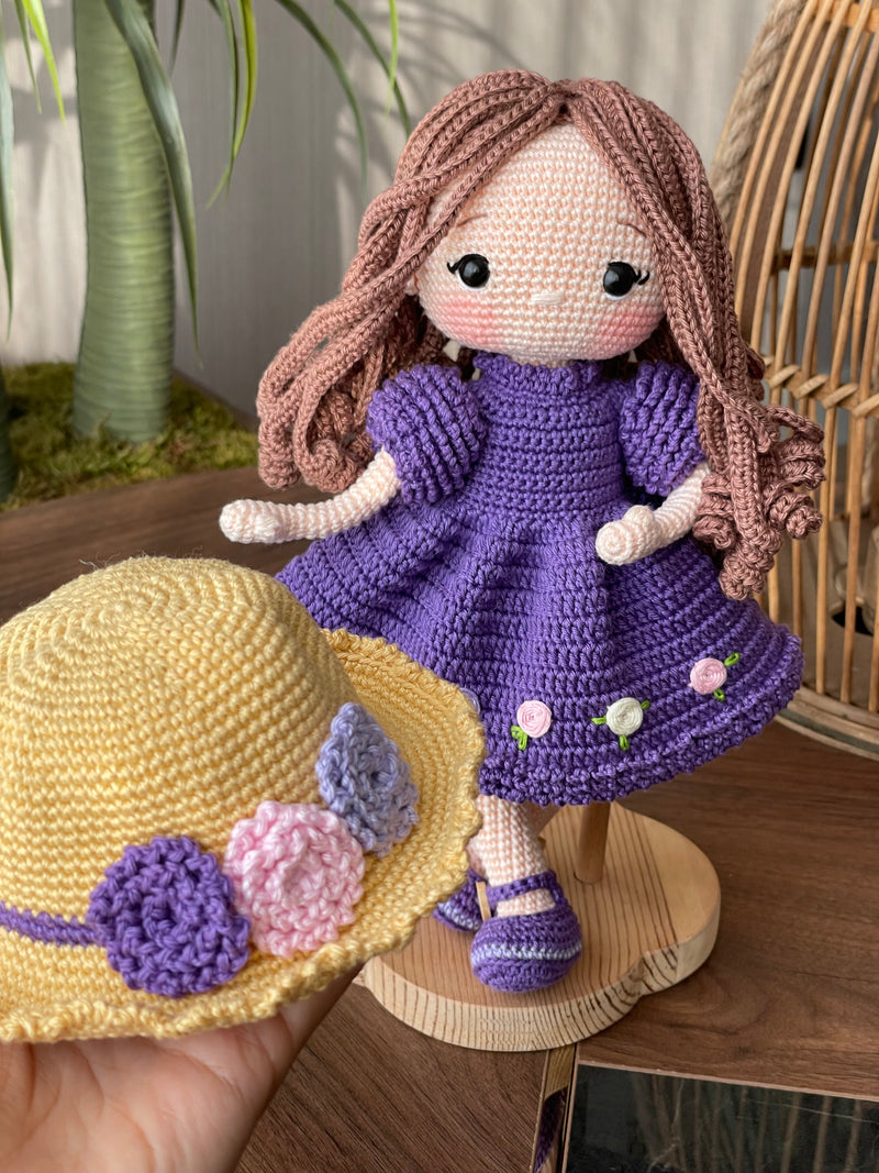 Handmade crocheted Purple Rose Girl Doll with long brown hair, wearing a purple dress with floral accents and a yellow hat. The doll is customizable with a name on a leather label, made using amigurumi techniques. Perfect gift for kids or room decor.