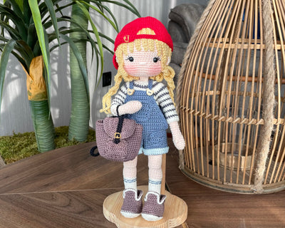 Handmade crocheted Mila Doll with a red cap, blue overalls, and a striped shirt, holding a small backpack, made with 100% cotton yarn and customizable with a name on a leather label, perfect for unique girl gifts and imaginative play.