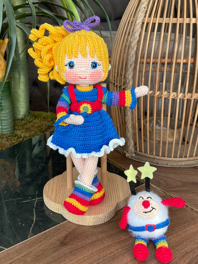 A handmade Rainbow Brite Girl Doll with vibrant yellow hair, holding Twink, a colorful star plush toy. The doll is crocheted using the amigurumi technique and personalized with a custom name on a leather label.