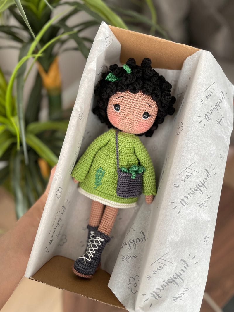 Handmade Rosemary dressed curly girl doll with a green outfit, crafted using amigurumi crochet technique, lying inside a gift box, personalized with a name on a leather label, ideal for gifts.