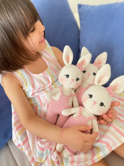 Pink color handmade crocheted Overall Girl Bunny Rabbit Toys with a cute bow and overalls held by a girl child. This amigurumi bunny doll is customizable with a name on a leather label, making it an ideal choice for nursery decor and personalized gifts.