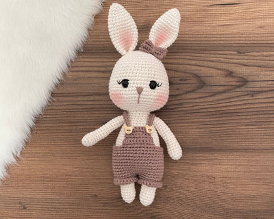 A mink color handmade crocheted Overall Girl Bunny Rabbit Toy with a cute bow and overalls. This amigurumi bunny doll is customizable with a name on a leather label, making it an ideal choice for nursery decor and personalized gifts.