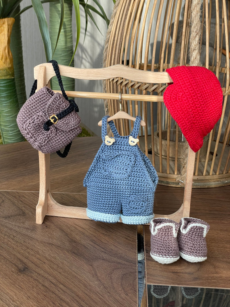 Handmade crocheted Mila Doll with a red cap, blue overalls, and a striped shirt, holding a small backpack, made with 100% cotton yarn and customizable with a name on a leather label.