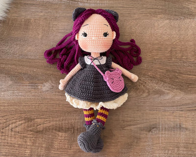 A handcrafted Cat Girl Doll with rich purple hair, wearing a dark dress with cat ears and holding a pink cat-shaped purse. The doll is crocheted using the amigurumi technique and personalized with a custom name on a leather label.