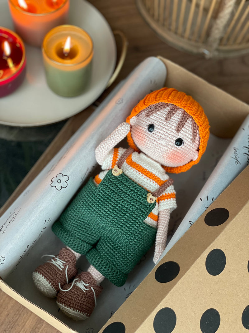 A handmade amigurumi crochet Boy With Pumpkin Hat, green overalls, and striped shirt. Lying inside a gift box. This customizable doll comes with a name label, perfect for fall or as an autumn gift.