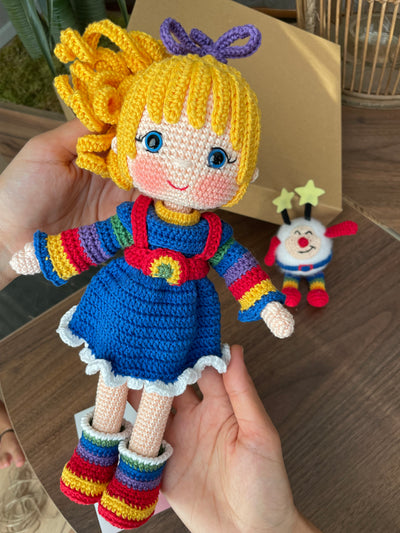 A handmade Rainbow Brite Girl Doll with vibrant yellow hair, holding Twink, a colorful star plush toy. The doll is crocheted using the amigurumi technique and personalized with a custom name on a leather label.