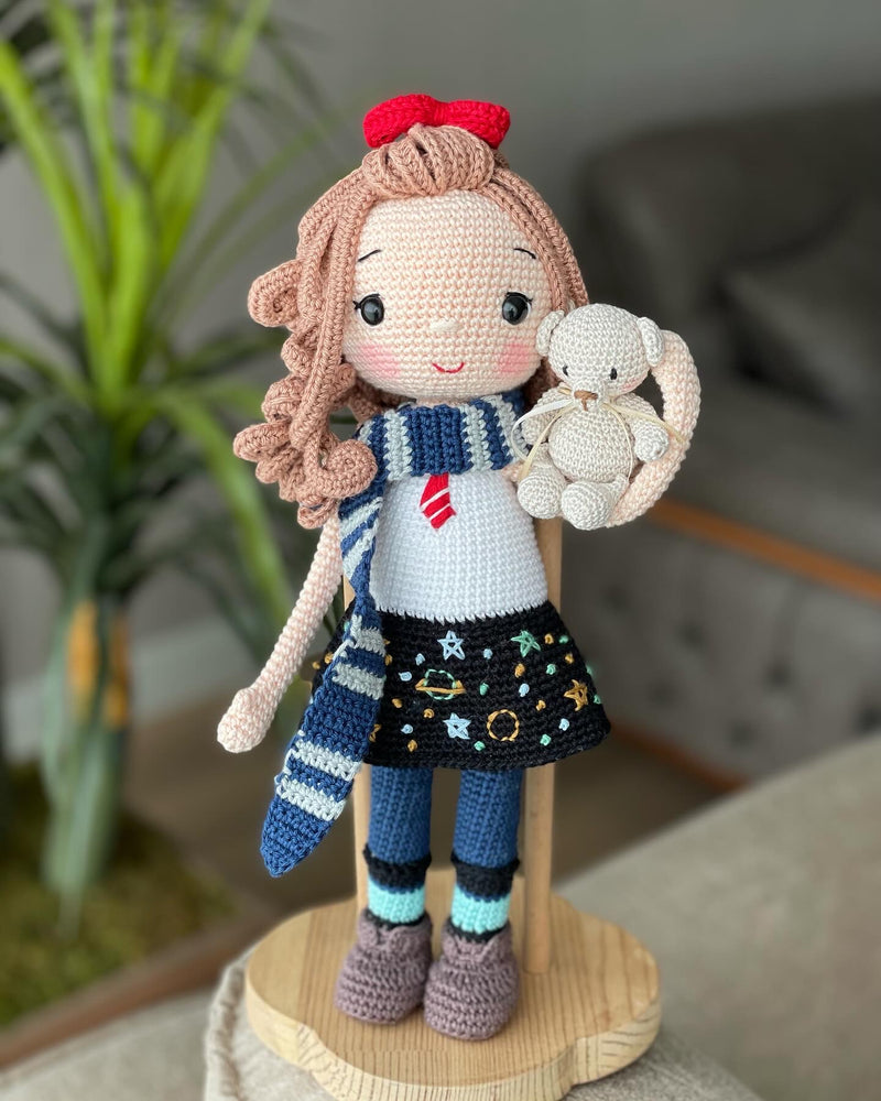 Handmade crochet Idealistic Girl Doll with a red coat, blue scarf, and holding a small teddy bear. This amigurumi doll is customizable with a name on a leather label, perfect for personalized gifts.