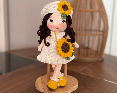 A handmade Sunflower Girl Doll with dark brown hair, wearing a white dress adorned with sunflowers, a matching sunflower beret, and yellow shoes, crocheted using the amigurumi technique and personalized with a custom name on a leather label.