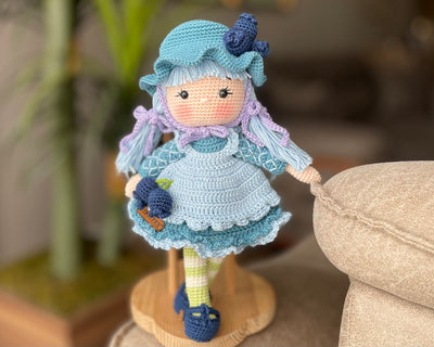 A handcrafted Blueberry Muffin Girl Doll featuring a detailed crochet blueberry accessory design, wearing a blue dress and hat, and personalized with a custom name on a leather label.