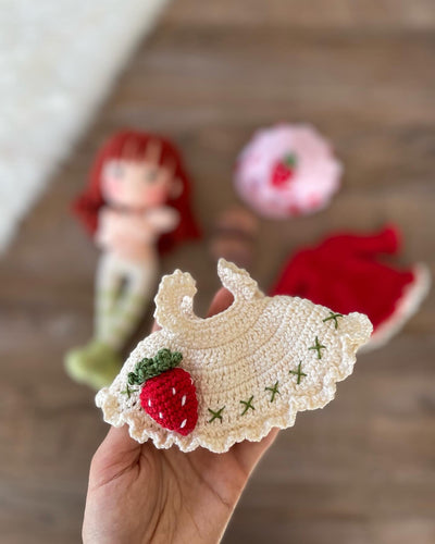 Handmade strawberry shortcake crochet doll with red hair, wearing a strawberry-themed outfit. Features a pink hat with red polka dots, red dress with white apron, and striped stockings. Perfect for children and collectors. Lying on a wooden surface.