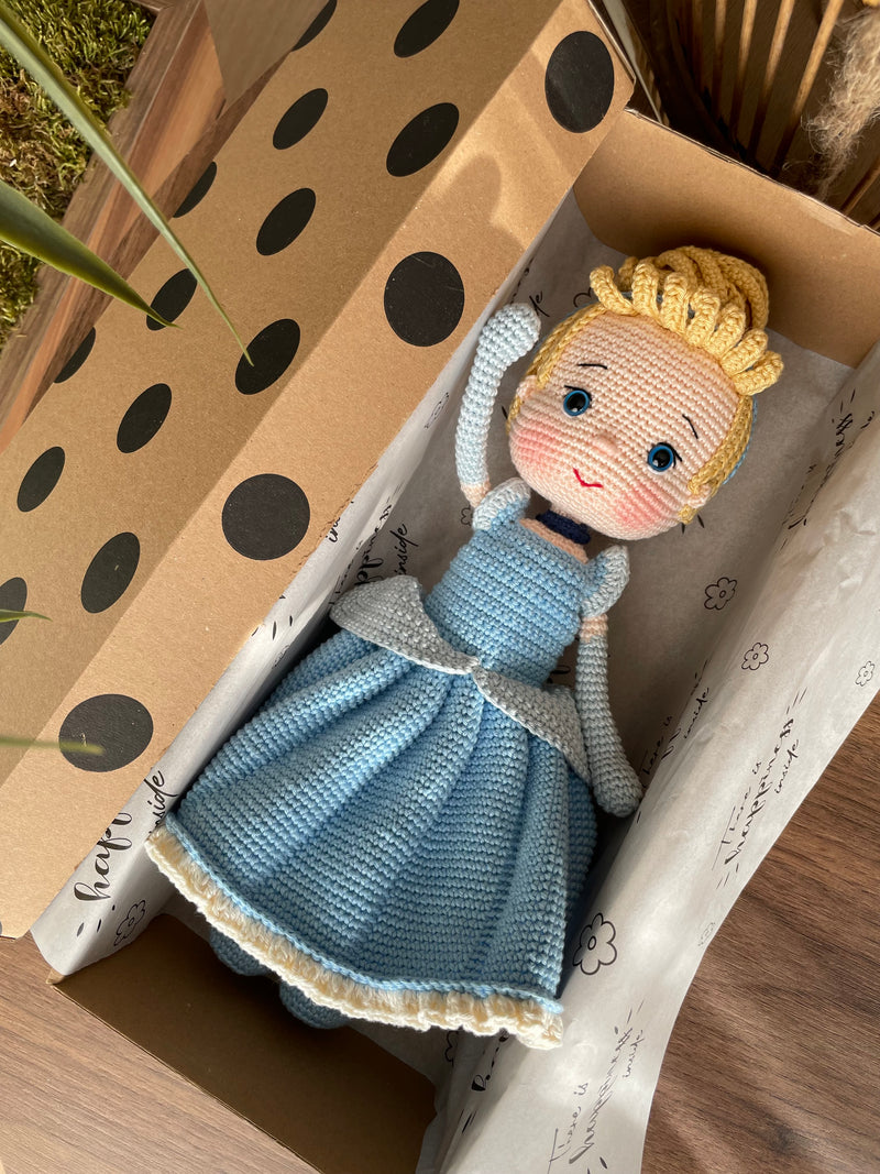 Handmade Cinderella Doll, crocheted using amigurumi technique with a blue dress, blonde hair, and custom name on a leather label, lying inside a gift box, perfect for princess lovers and gifts.