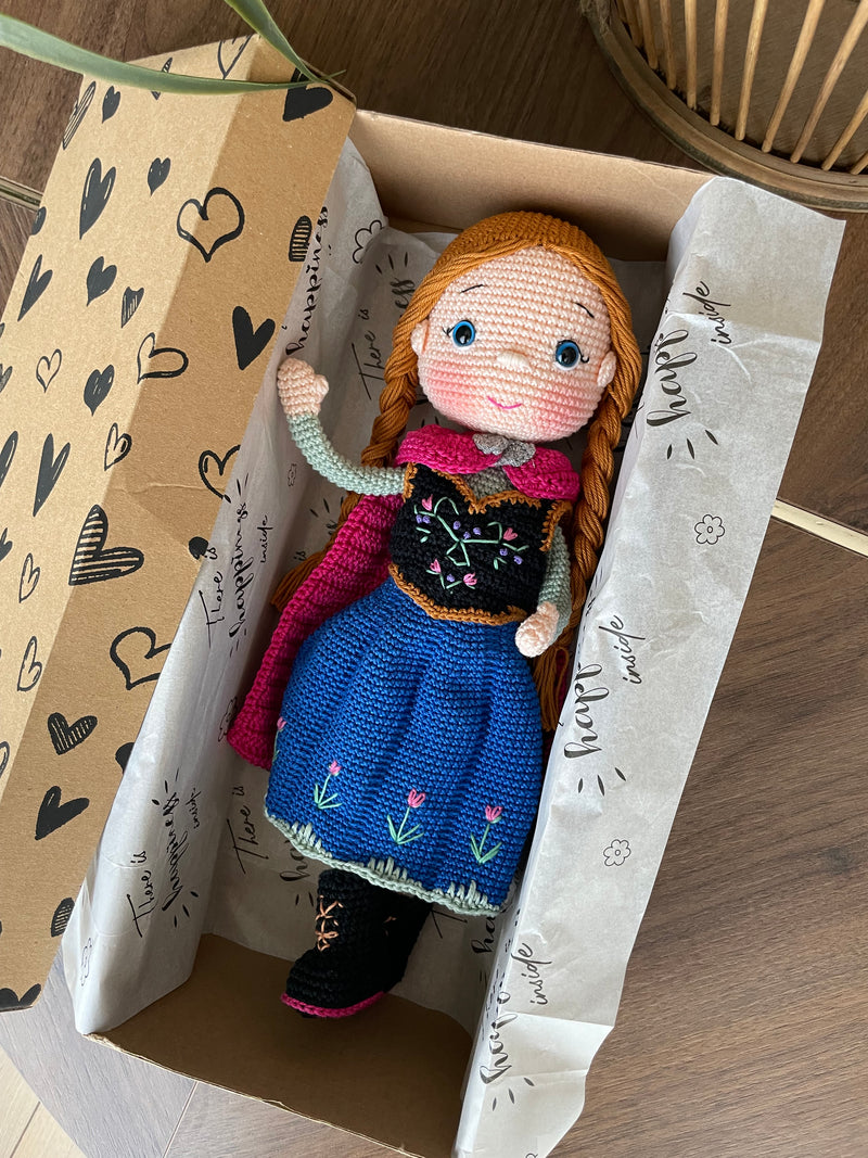 Handmade crocheted amigurumi dolls of Elsa and Anna, inspired by Frozen. Detailed with colorful outfits, perfect for custom name personalization or gifting. Lying inside a gift box.