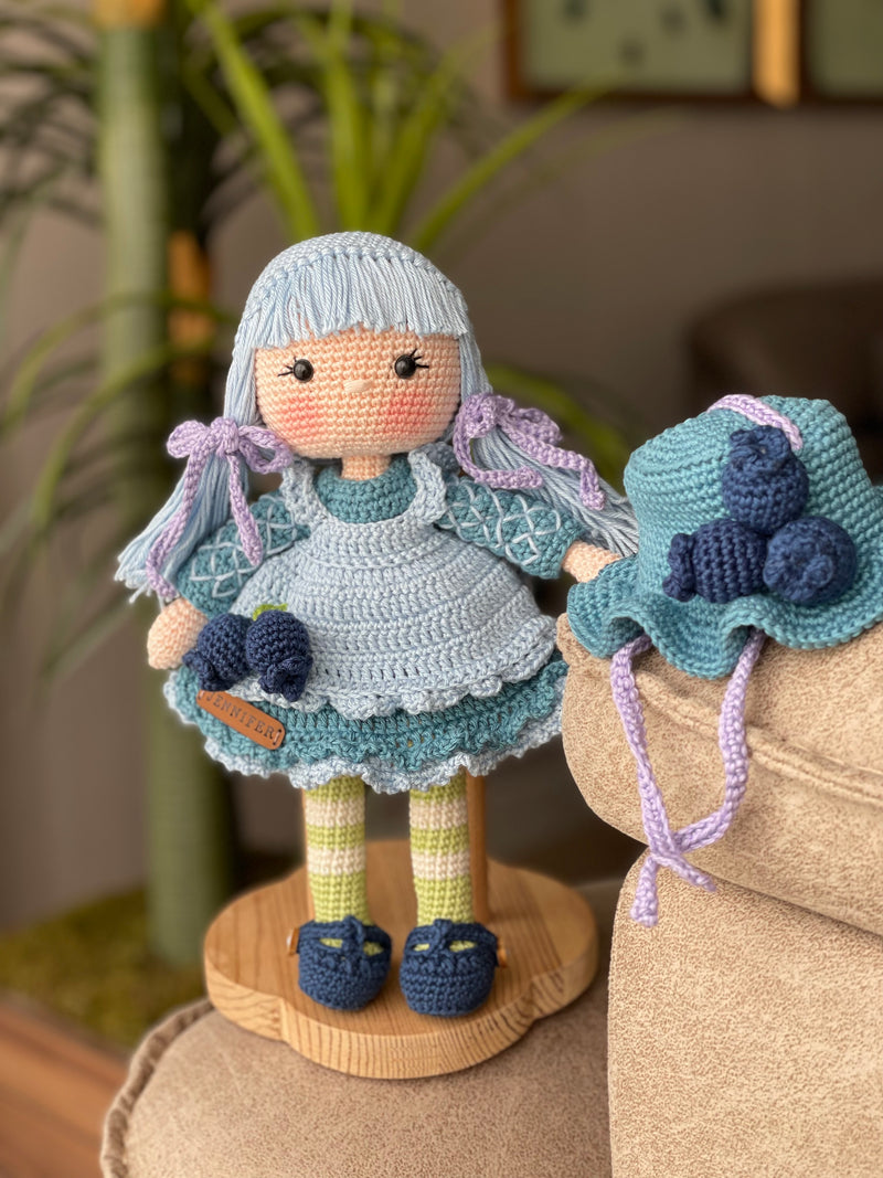 A handcrafted Blueberry Muffin Girl Doll featuring a detailed crochet blueberry accessory design, wearing a blue dress and hat, and personalized with a custom name on a leather label.