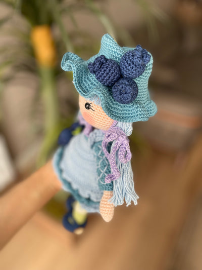 A handcrafted Blueberry Muffin Girl Doll featuring a detailed crochet blueberry accessory design, wearing a blue dress and hat, and personalized with a custom name on a leather label.