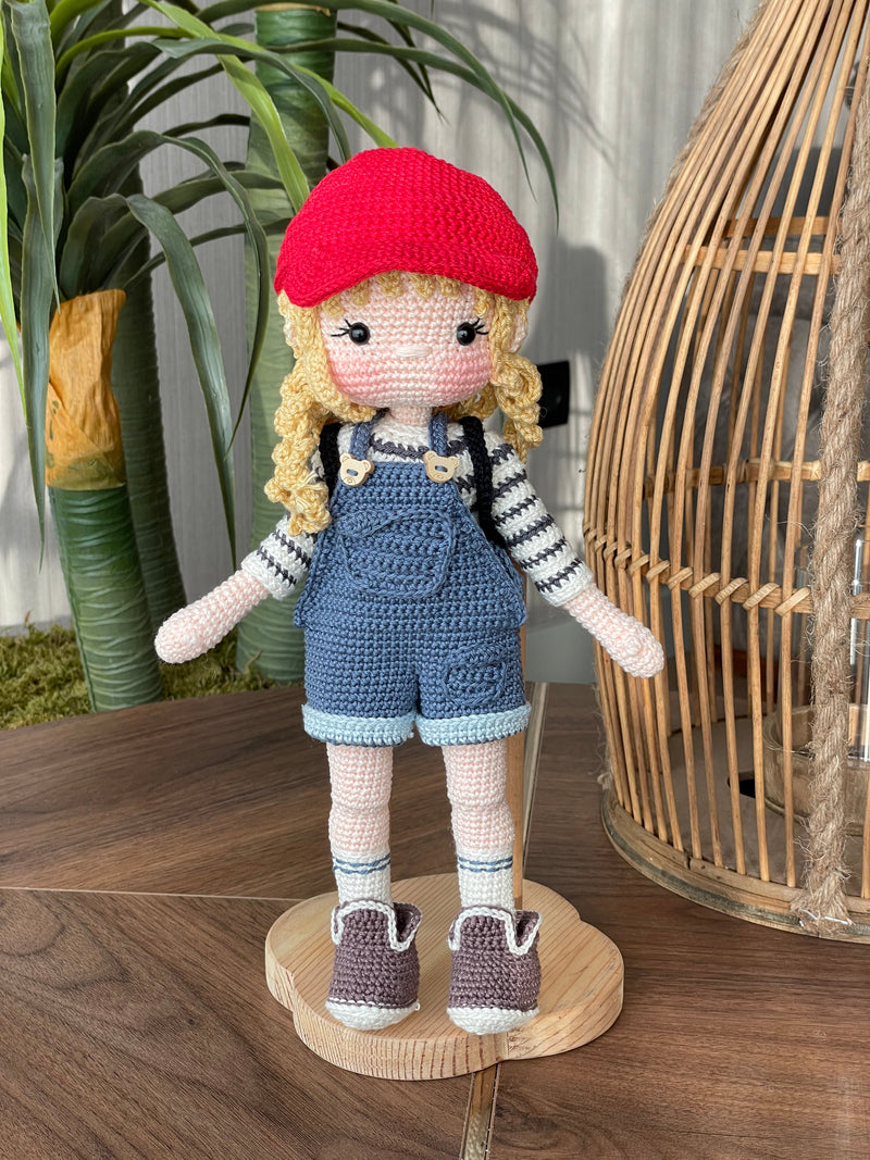 Handmade crocheted Mila Doll with a red cap, blue overalls, and a striped shirt, holding a small backpack, made with 100% cotton yarn and customizable with a name on a leather label.