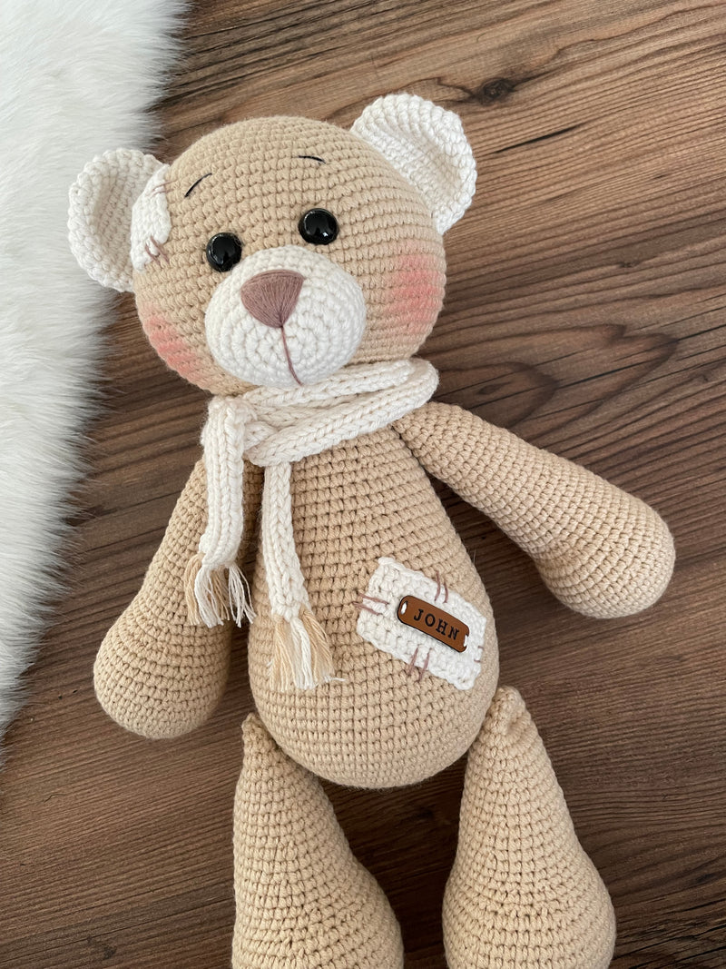 A handmade beige teddy bear toy crafted with the amigurumi technique, featuring a soft texture, a personalized leather name patch, and a cute, cuddly design.