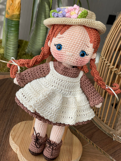 Handmade crocheted Anne Shirley Doll with long braided hair, wearing a white apron and a floral hat. The doll is customizable with a name on a leather label, perfect as a personalized gift or room decor.