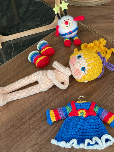 A handmade Rainbow Brite Girl Doll with vibrant yellow hair, holding Twink, a colorful star plush toy. The doll is crocheted using the amigurumi technique and personalized with a custom name on a leather label.