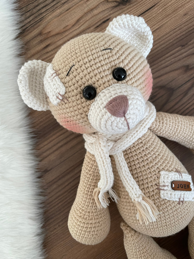 A handmade beige teddy bear toy crafted with the amigurumi technique, featuring a soft texture, a personalized leather name patch, and a cute, cuddly design.