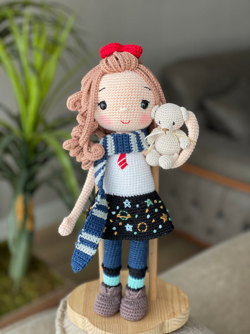 Handmade crochet Idealistic Girl Doll with a red coat, blue scarf, and holding a small teddy bear. This amigurumi doll is customizable with a name on a leather label, perfect for personalized gifts.