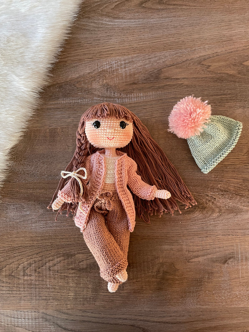 A handcrafted Pink Cardigan Cool Girl Doll with long brown hair, wearing a stylish pink cardigan and beanie, crocheted using the amigurumi technique, and personalized with a custom name on a leather label.