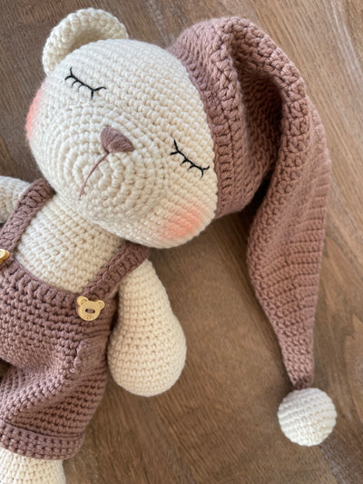 Handmade crocheted Sleeping Teddy Bear Toy in soft, neutral tones with a cozy hat and overalls, customizable with a name on a leather label. Perfect for bedtime cuddles and nursery decor.