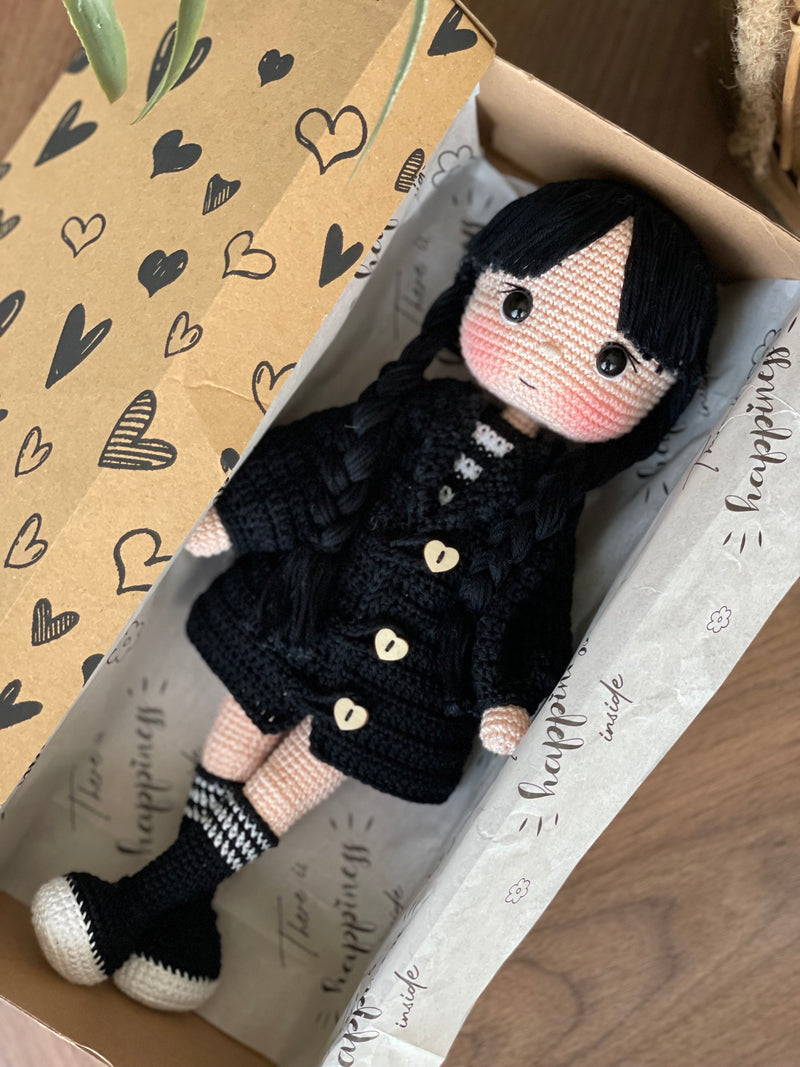 Handmade Wednesday Addams Doll with black braids, a black dress, and a spooky themed accessory, crocheted using the amigurumi technique and personalized with a name. Lying inside a gift box. Perfect for kids and Halloween Gift.