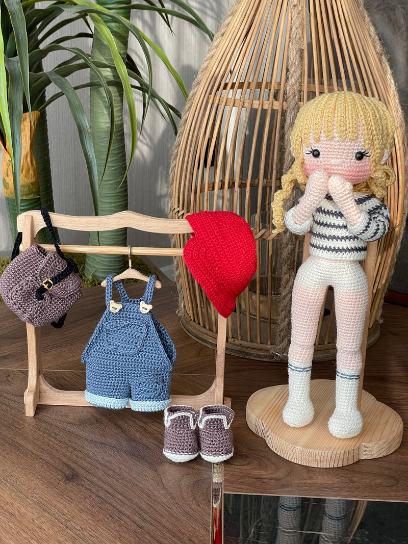 Handmade crocheted Mila Doll with a red cap, blue overalls, and a striped shirt, holding a small backpack, made with 100% cotton yarn and customizable with a name on a leather label.