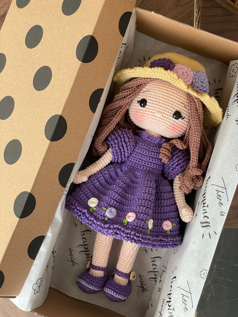 Handmade crocheted Purple Rose Girl Doll with long brown hair, wearing a purple dress with floral accents and a yellow hat. The doll is customizable with a name on a leather label, made using amigurumi techniques. Lying inside a gift box. Perfect gift for