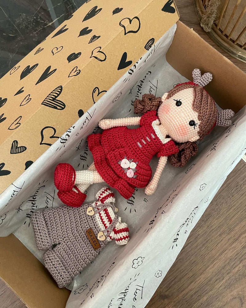 Handmade crochet Forest Girl Doll with two interchangeable dresses and matching accessories. This amigurumi doll is customizable with a name on a leather label, perfect for personalized gifts. Lying inside a gift box.