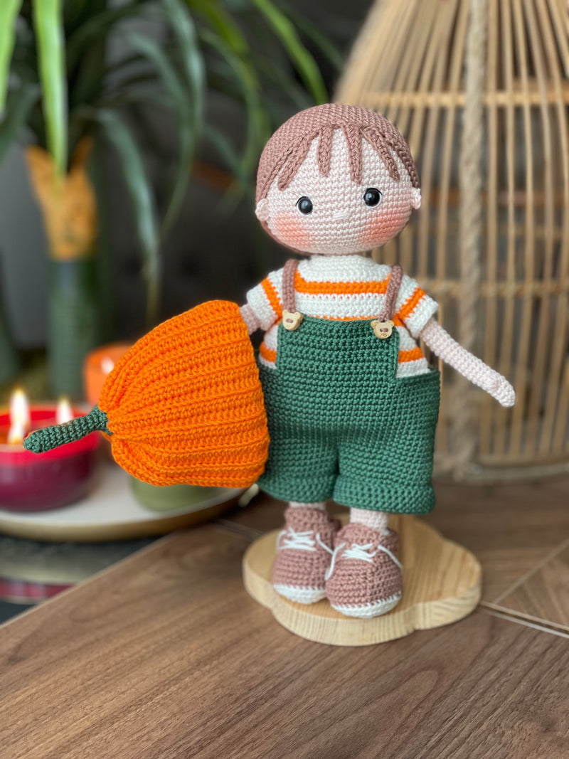 A handmade amigurumi crochet Boy With Pumpkin Hat, green overalls, and striped shirt. This customizable doll comes with a name label, perfect for fall or as an autumn gift.