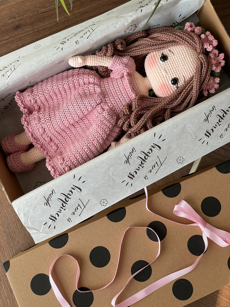 Handmade pink princess girl doll, crocheted using amigurumi technique. Features a beautiful dress and flower crown, customizable with a name on a leather label. Lying inside a gift box. Perfect for kids and collectors.