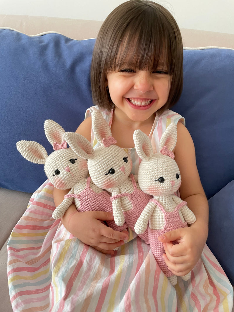 Pink color handmade crocheted Overall Girl Bunny Rabbit Toys with a cute bow and overalls held by a girl child. This amigurumi bunny doll is customizable with a name on a leather label, making it an ideal choice for nursery decor and personalized gifts.