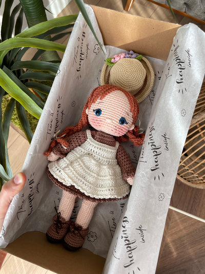 Handmade crocheted Anne Shirley Doll with long braided hair, wearing a white apron and a floral hat. Lying inside a gift box. The doll is customizable with a name on a leather label, perfect as a personalized gift or room decor.