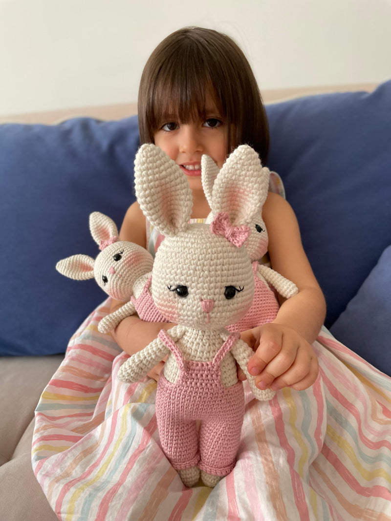 Pink color handmade crocheted Overall Girl Bunny Rabbit Toys with a cute bow and overalls held by a girl child. This amigurumi bunny doll is customizable with a name on a leather label, making it an ideal choice for nursery decor and personalized gifts.