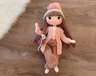 A handcrafted Pink Cardigan Cool Girl Doll with long brown hair, wearing a stylish pink cardigan and beanie, crocheted using the amigurumi technique, and personalized with a custom name on a leather label.