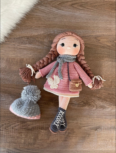 Handmade Pink Winter Girl Doll in a pink coat, gray scarf, and hat with braids, made with 100% cotton yarn, customizable with a name on a leather label.