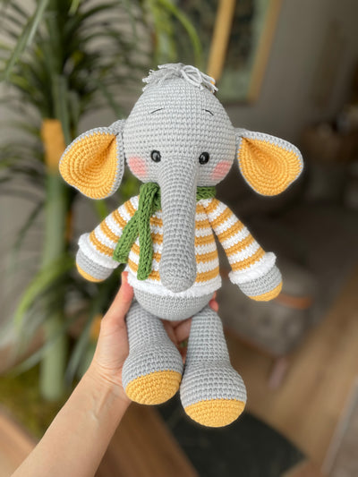 A handmade crochet elephant toy featuring a soft, cuddly design with large yellow ears, yellow striped sweater, and a green scarf, made using the amigurumi technique and personalized with a custom name on a leather label.