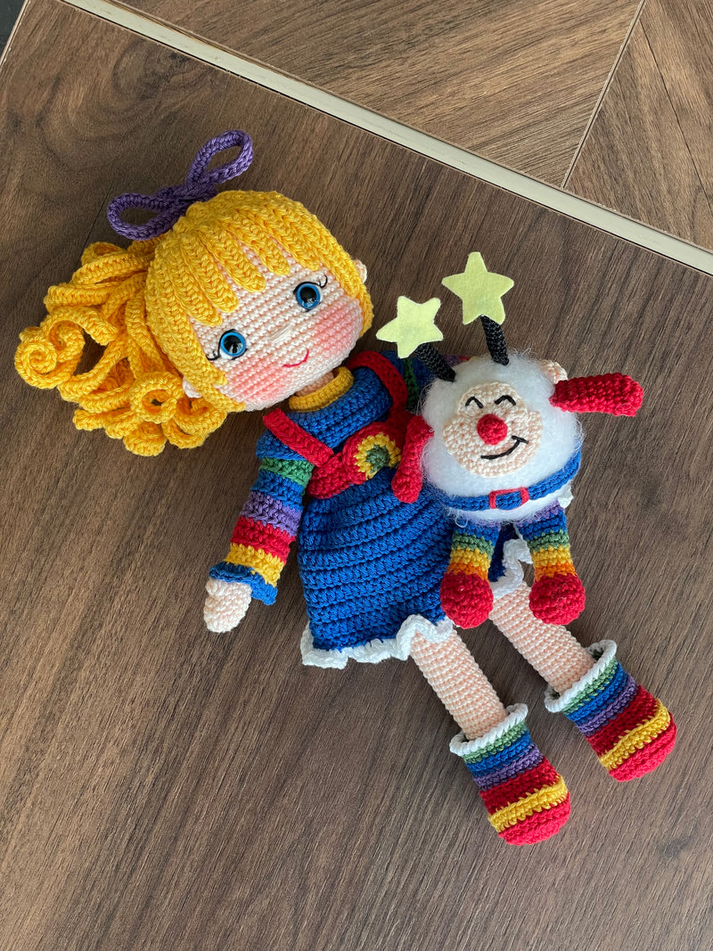 A handmade Rainbow Brite Girl Doll with vibrant yellow hair, holding Twink, a colorful star plush toy. The doll is crocheted using the amigurumi technique and personalized with a custom name on a leather label.