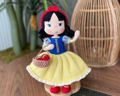 Handmade Snow White Doll wearing a classic yellow dress with a blue bodice, holding a basket of apples, made with 100% cotton yarn, customizable with a name on a leather label.