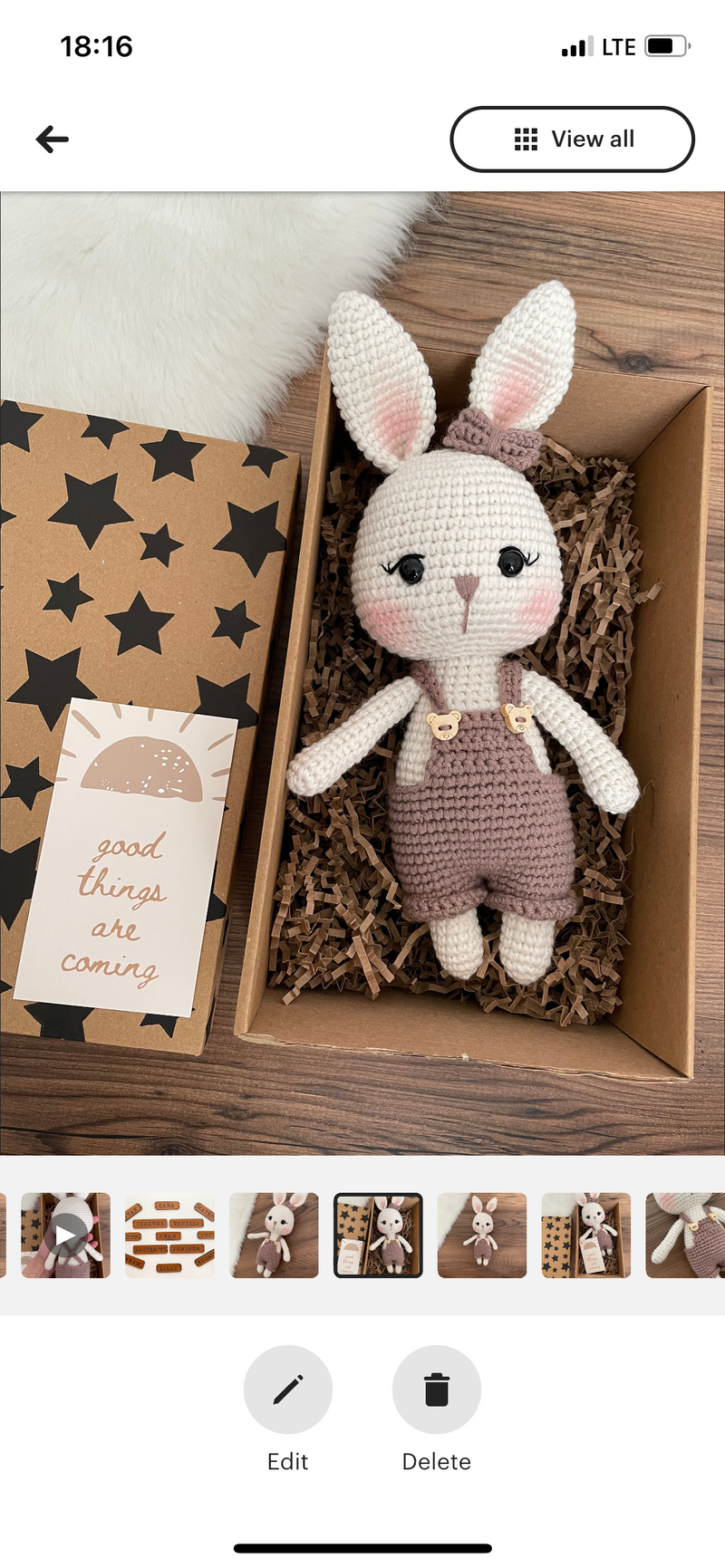 A mink color handmade crocheted Overall Girl Bunny Rabbit Toy with a cute bow and overalls, lying inside a gift box. This amigurumi bunny doll is customizable with a name on a leather label, making it an ideal choice for nursery decor and personalized gif