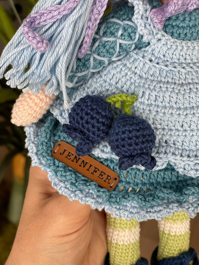 A handcrafted Blueberry Muffin Girl Doll featuring a detailed crochet blueberry accessory design, wearing a blue dress and hat, and personalized with a custom name on a leather label.