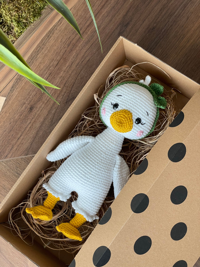 Handmade crocheted Duck Toy with a yellow beak and feet, customizable with a name on a leather label, crafted using amigurumi techniques. Lying inside a gift box. A perfect gift for kids or as nursery decor.